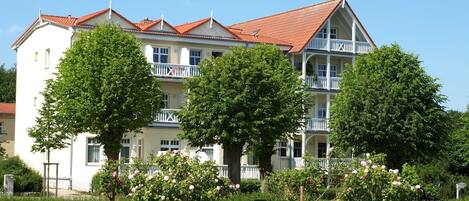 A multi-story building with a red-tiled roof, white balconies, and lush greenery in the foreground.