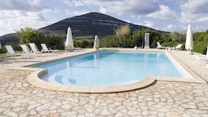 A swimming pool with lounge chairs and umbrellas, surrounded by stone flooring, with a mountain in the background. - Holiday apartment with air conditioning (Alghero)