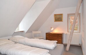 A bedroom with a sloped ceiling, a wooden dresser, a bedside table with a lamp, a chair, and a window with a view of trees.