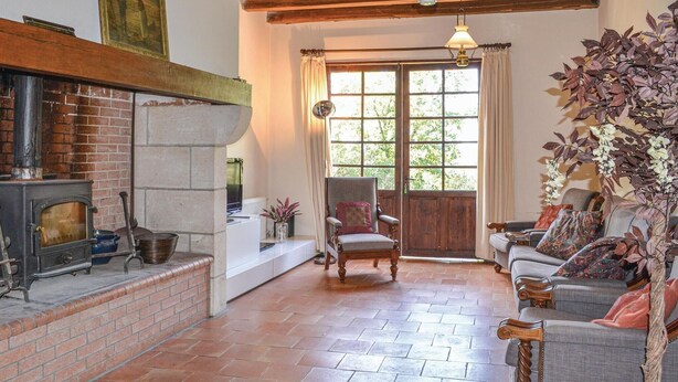 A cozy living room with a fireplace, a comfortable armchair, a sofa set, and a large window with a view of greenery.