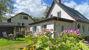 Une maison à l’extérieur blanc, avec un toit en pente et une cheminée bien visible. Au premier plan, on aperçoit un jardin avec des fleurs jaunes et violettes.