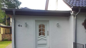 Une maison blanche avec le numéro « 18 » sur la porte, un petit porche avec des marches et deux plantes en pot ornées de fleurs roses.