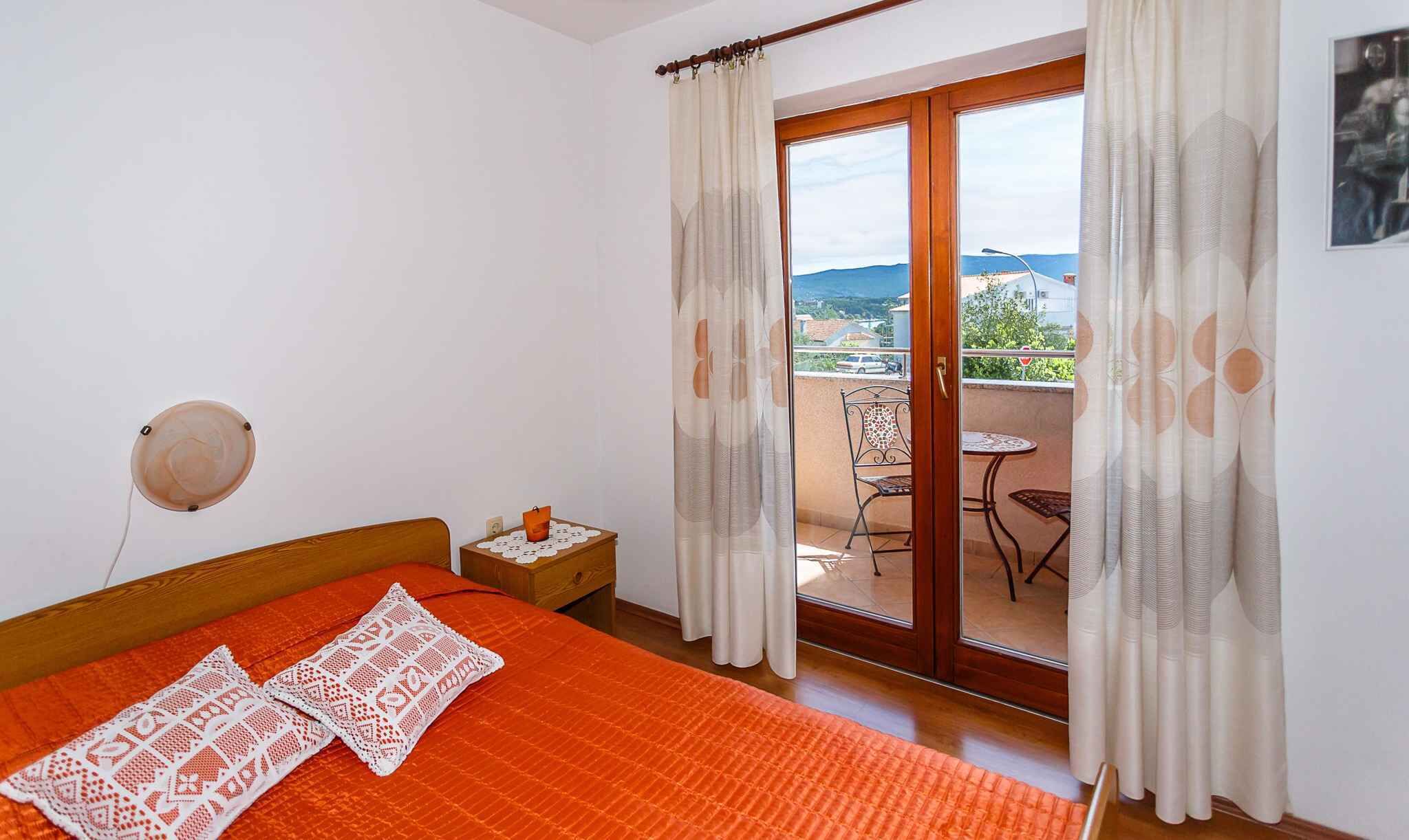 A bedroom with a wooden bed, an orange bedspread, a white crocheted pillow, a nightstand with a candle, and a balcony with a table and chairs.