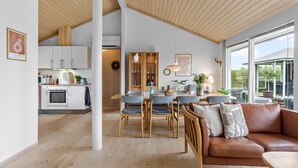A spacious living area with a wooden ceiling, a dining table with chairs, a brown leather sofa, and a large window offering a view of the outdoors. - Cozy home in Nakskov with kitchen (Nakskov)