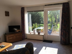 Una sala de estar con un televisor sobre un mueble de madera, un sofá, una mesa de centro con un control remoto y vistas a un jardín a través de una puerta corrediza de vidrio.