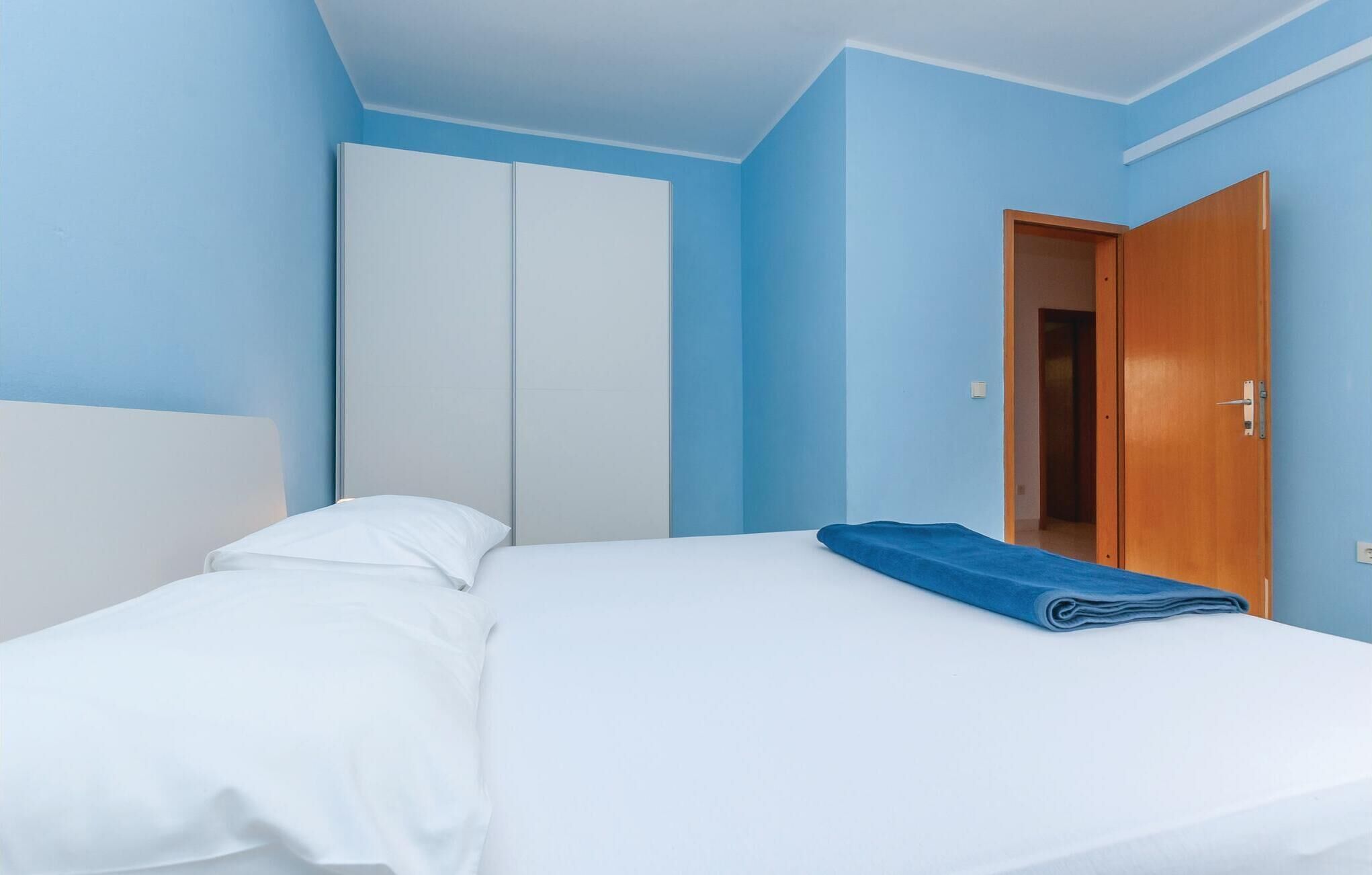 A bedroom with a bed, white bedding, a blue wall, a wardrobe, and a wooden door.