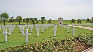 A cemetery with numerous white crosses arranged in rows, a central monument, and a pathway leading to it.