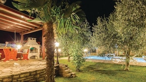 Un espacio al aire libre nocturno con una piscina de piedra, palmeras y una zona de estar cubierta con chimenea de piedra.