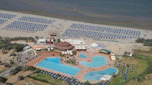 An aerial view of a resort with multiple swimming pools, beach areas with umbrellas, and a variety of buildings including a large hotel structure.