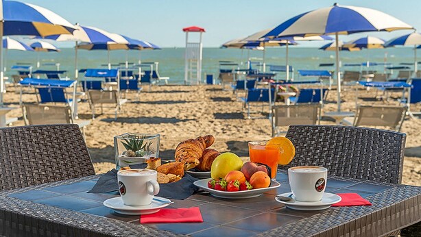 A beachside breakfast table with croissants, fruit, and coffee, overlooking the beach with umbrellas and chairs.
