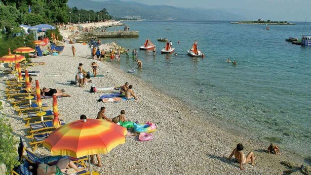 Een kiezelstrand met ligbedden en parasols, boten op het water, en mensen die zwemmen.