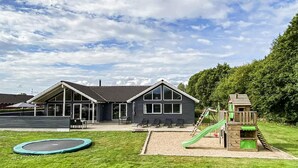 Un edificio de dos pisos con ventanas amplias y techo plano, un trampolín y un parque infantil con tobogán.