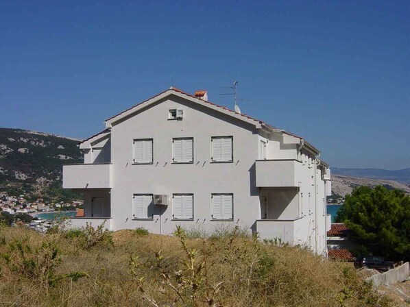 A two-story building with a flat roof and multiple windows, surrounded by natural landscape and a body of water.