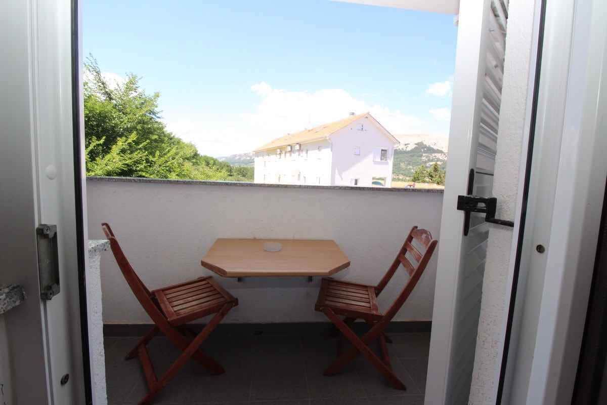 A balcony with a table and two wooden chairs, overlooking a white building and greenery.