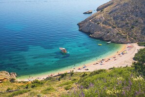Una playa costera con agua turquesa cristalina, acantilados rocosos y una orilla de arena con sombrillas y gente.