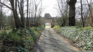 Ein schmaler, unbefestigter Weg führt zu einem steinernen Torbogen, im Hintergrund ist ein Gebäude zu sehen.