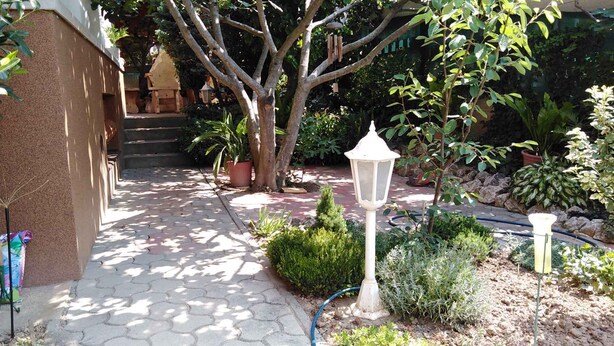 A garden pathway leading to a building entrance, surrounded by potted plants and a white lamp post.