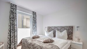 A bedroom with a large bed, white bedding, patterned curtains, and a nightstand with a lamp. - Villa Margarete Whg. 6 - Villa Margarete, BEACH NEXT (Heringsdorf)