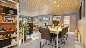 Un espace repas moderne avec un plafond en bois, une table centrale entourée de chaises et une bibliothèque remplie de divers objets.