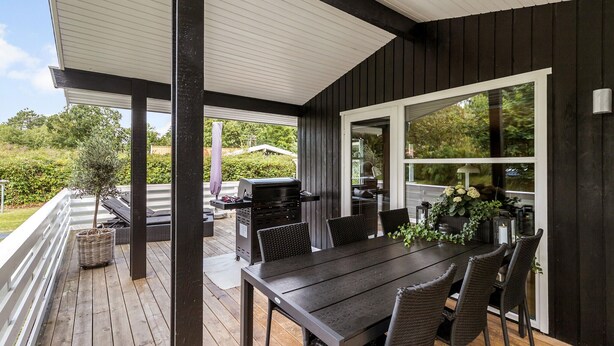 Une terrasse extérieure couverte, équipée d’une table et de chaises, d’un barbecue et offrant une vue sur la verdure.