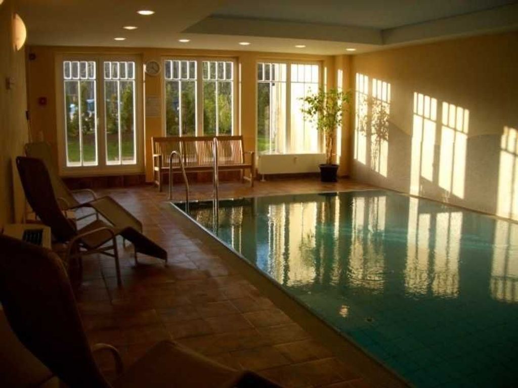 An indoor swimming pool with a bench and chairs along the side, large windows, and a potted plant.