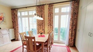 A dining area with a table and chairs, a red rug, and a view through large windows. - (109) 2-room apartment - apartment house Dünenschloss (Kühlungsborn)