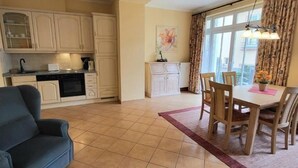 A spacious living area with a dining table, a sofa, and a kitchen area with cabinets and a stove. - (109) 2-room apartment - apartment house Dünenschloss (Kühlungsborn)