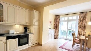 A kitchen with white cabinets, a stove, and a dining area with a table and chairs. - (109) 2-room apartment - apartment house Dünenschloss (Kühlungsborn)
