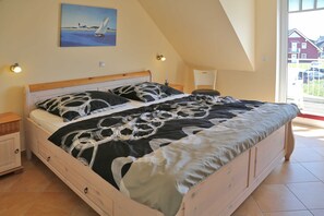 A bedroom with a wooden bed, a painting of a sailboat, and a view of houses through the window.