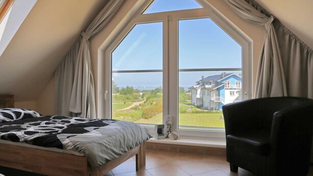 Ein Schlafzimmer mit großem Fenster, einem Bett, einem Sessel und Blick auf ein blaues Haus.