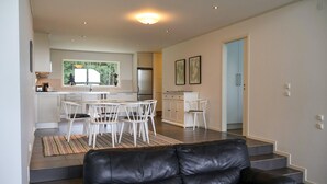 Una sala moderna con un sofá de cuero negro, un comedor con sillas blancas y una cocina con armarios blancos y un refrigerador.