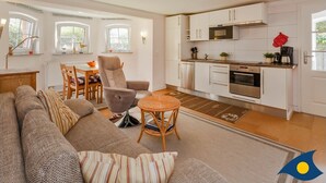 A modern living room with a sofa, armchair, and small table. Adjacent is a kitchen with white cabinets, a microwave, and an oven. There are windows with curtains and a ceiling light fixture. - Villa Exss Whg. 01 /- - VE 01 (Heringsdorf)