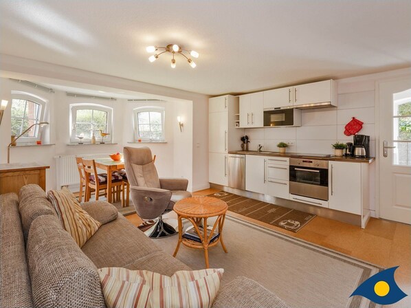 A modern living room with a sofa, armchair, and small table. Adjacent is a kitchen with white cabinets, a microwave, and an oven. There are windows with curtains and a ceiling light fixture.