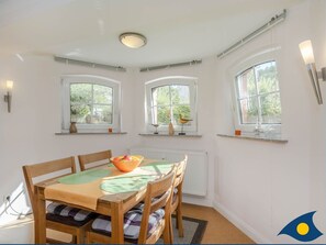 A dining area with a wooden table, chairs, and a bowl of fruit. There are three windows with shelves above them.