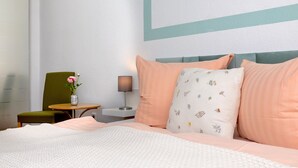 Une chambre d’hôtel bien rangée, avec un lit, une chaise, une petite table sur laquelle est posé un vase de fleurs et une lampe.