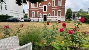 Un jardin où s’épanouissent des rosiers et où se trouve un fauteuil en osier.
