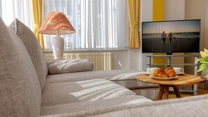 Un salon avec un canapé, une table basse sur laquelle sont posés des croissants, un téléviseur diffusant une scène de plage et une lampe.
