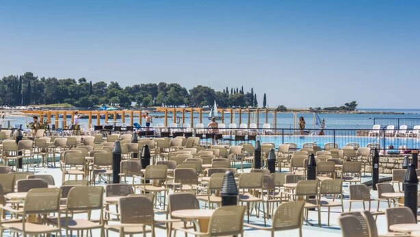 A poolside area with numerous chairs and tables, a clear view of the sea, and a distant shoreline with trees and buildings. - Studio with outdoor pool (Umag)