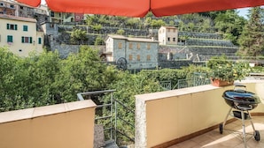 A balcony with a red umbrella, a grill, and a view of a hillside with buildings.
