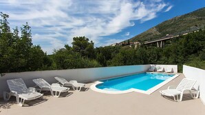 Una piscina con lettini bianchi, circondata da vegetazione e con montagne sullo sfondo.