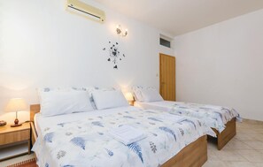 Uma cama de casal com colcha estampada em folhas, um abajur, ar condicionado fixado na parede e uma porta de madeira.