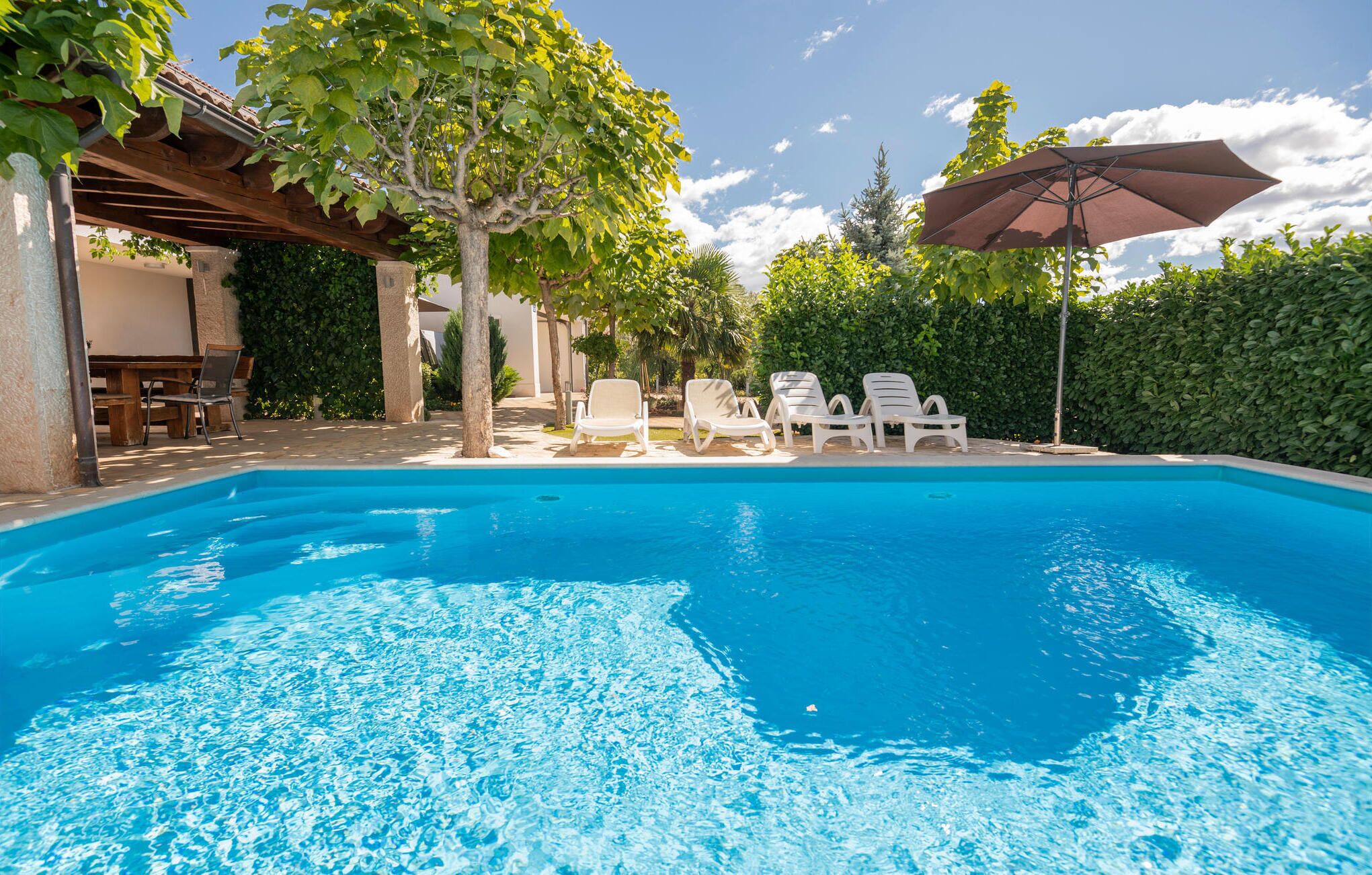 A clear blue swimming pool with white lounge chairs and a pergola with a table and chairs.
