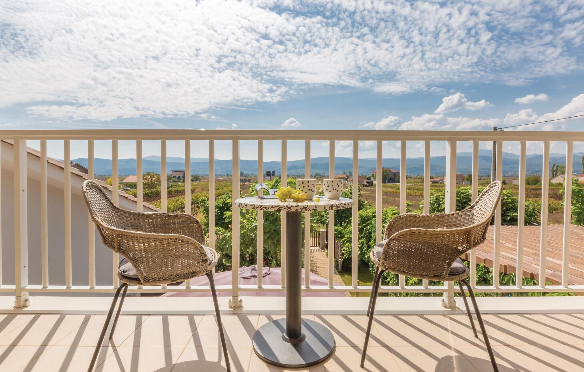 Ein Balkon mit zwei Korbstühlen, einem kleinen runden Tisch mit Blumenvase und Blick auf Weinberge und Berge.
