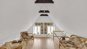 Una habitación tipo loft con techos altos, techo inclinado y un ventanal grande. Hay dos sillones de mimbre con mantas de piel, una mesa de centro de vidrio y una pequeña mesa de madera con una bandeja. Las paredes son blancas y hay cuadros enmarcados.