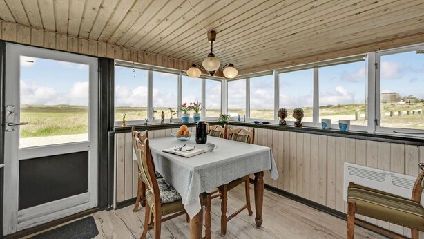 Un comedor con una mesa lista para dos, sillas de madera, vista a la playa a través de grandes ventanales y un techo con vigas de madera.