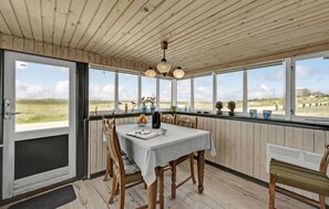 Un comedor con una mesa lista para dos, sillas de madera, vista a la playa a través de grandes ventanales y un techo con vigas de madera.