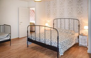 Un dormitorio con estructura de cama metálica, ropa de cama estampada, una mesita de noche con lámpara y un armario.