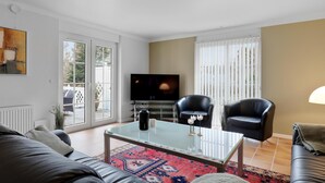 A modern living room with a glass coffee table, two leather armchairs, a sofa, a flat-screen TV, and a large window with blinds.