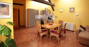 A dining area with a wooden table and wicker chairs, a kitchen with modern appliances, and a yellow wall with a clock and a framed picture.