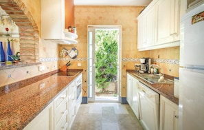 Cocina con encimeras de granito, armarios blancos, cafetera y vista a la vegetación a través de la puerta.